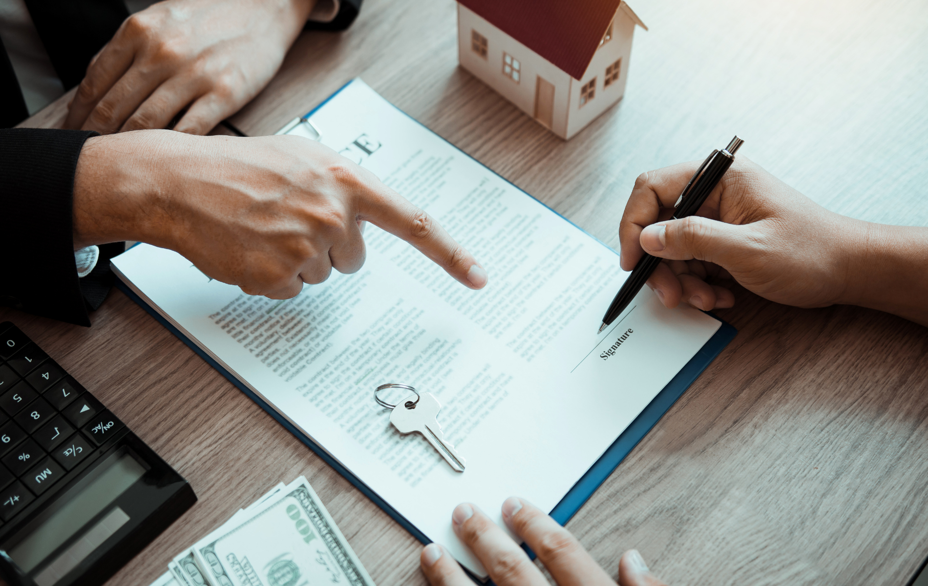 A person points to a contract on a wooden table while another signs it near a house model, keys, calculator, and cash.