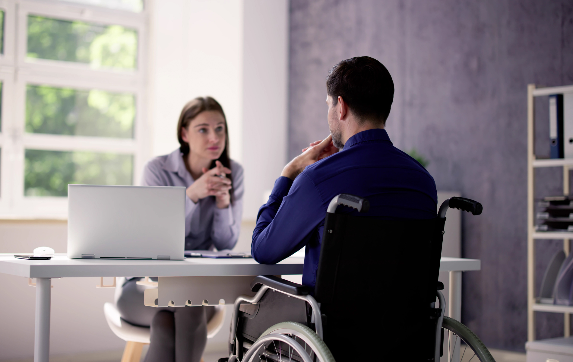 A professional sitting at a desk faces a person in a wheelchair during a meeting in a brightly lit office.