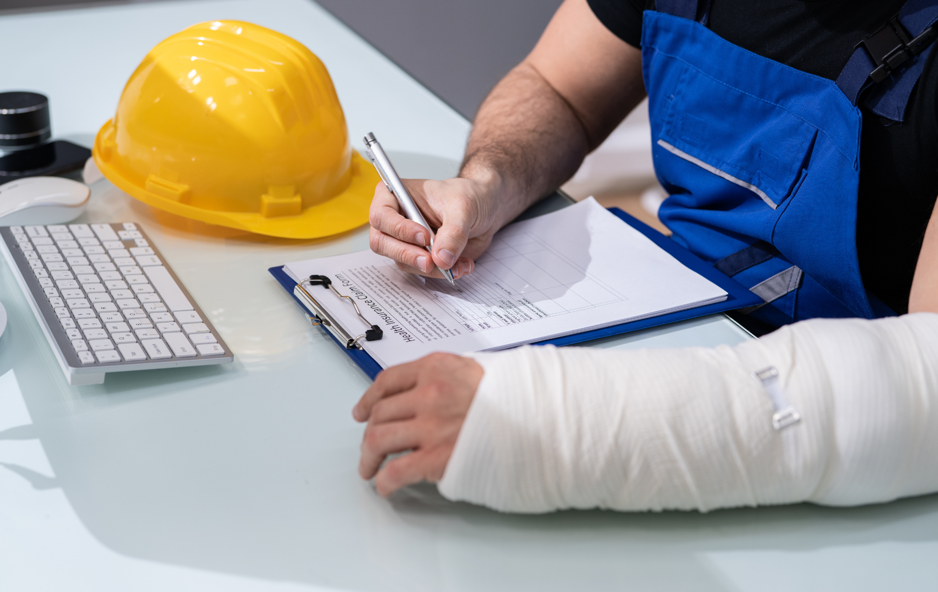 A person in a blue uniform with a cast on their arm writes on a clipboard at a desk with a yellow hard hat and keyboard.