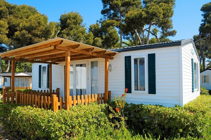 A white mobile home with dark green shutters and a wooden porch, set among trees and green shrubs under a clear blue sky.