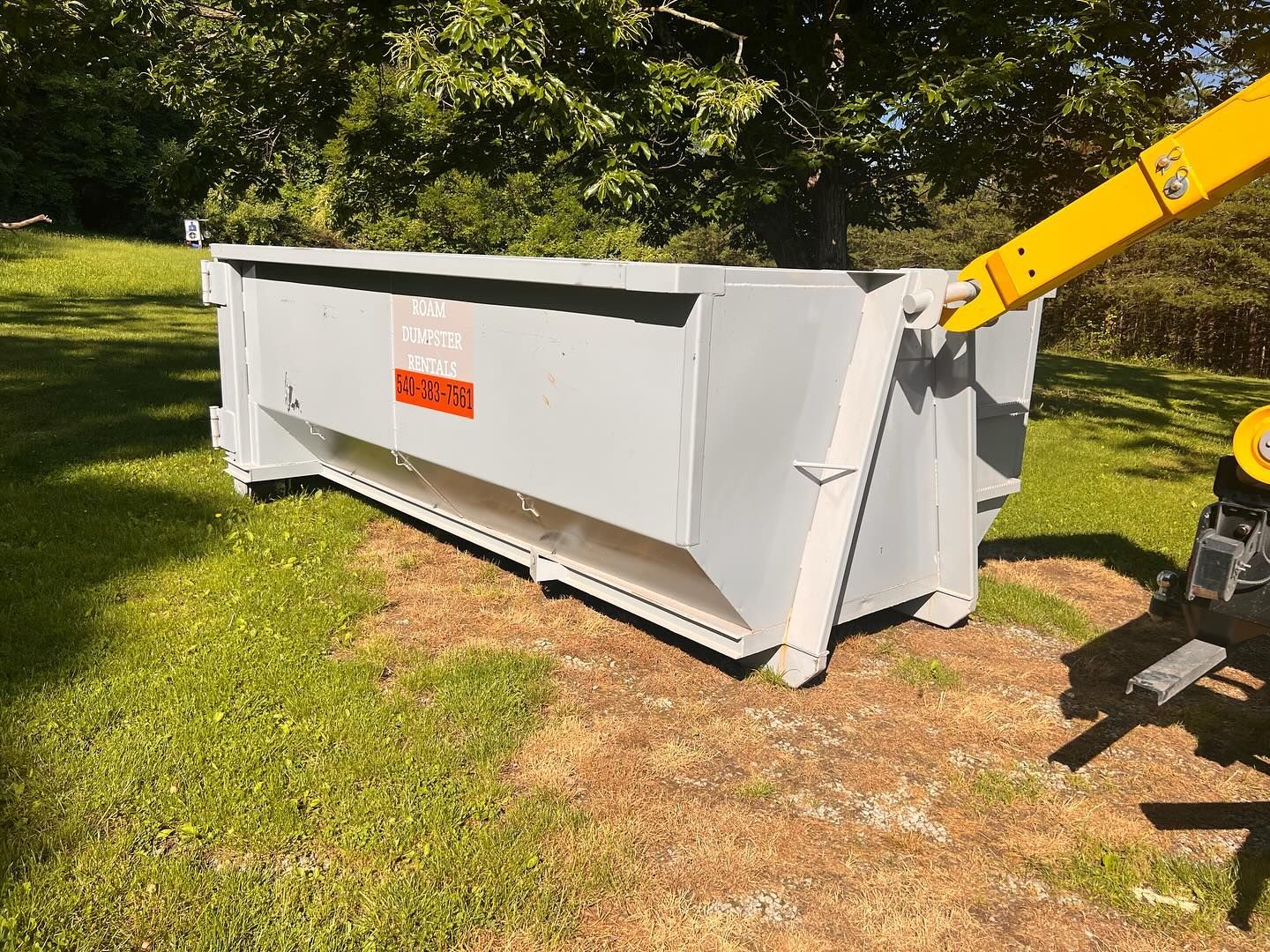 A large white dumpster is sitting in the grass next to a yellow crane.