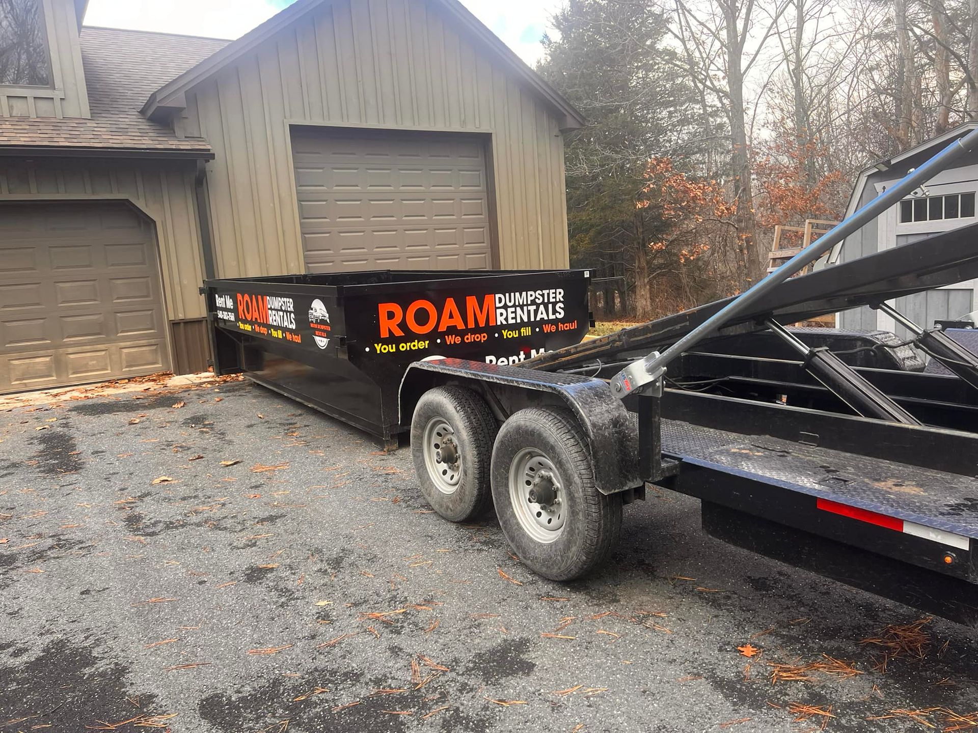 A dumpster is being towed by a trailer in front of a garage.