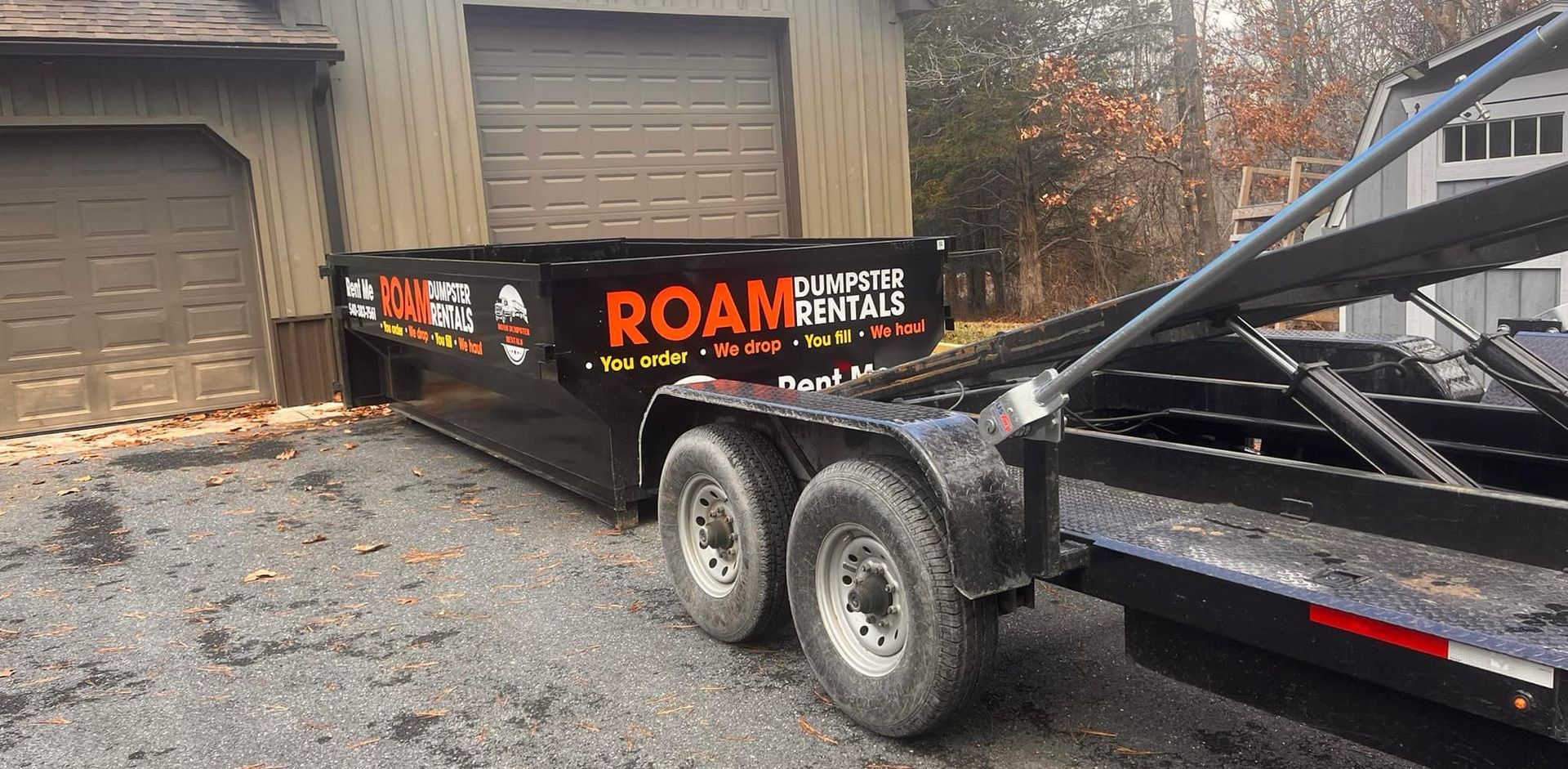 A dumpster on a trailer is parked in front of a garage.