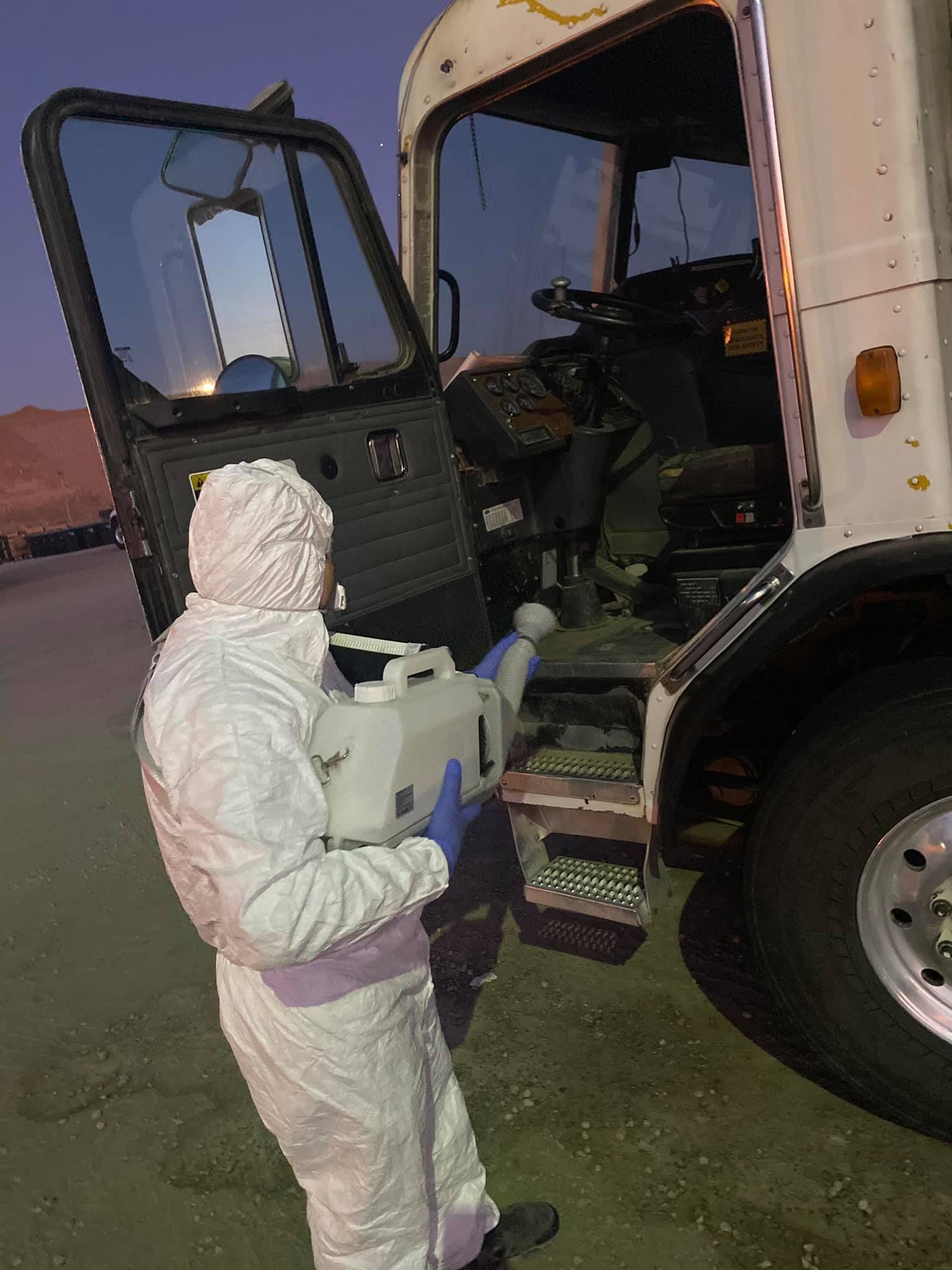 A person in a white protective suit uses a handheld sprayer to disinfect the interior of a truck cab at dusk.