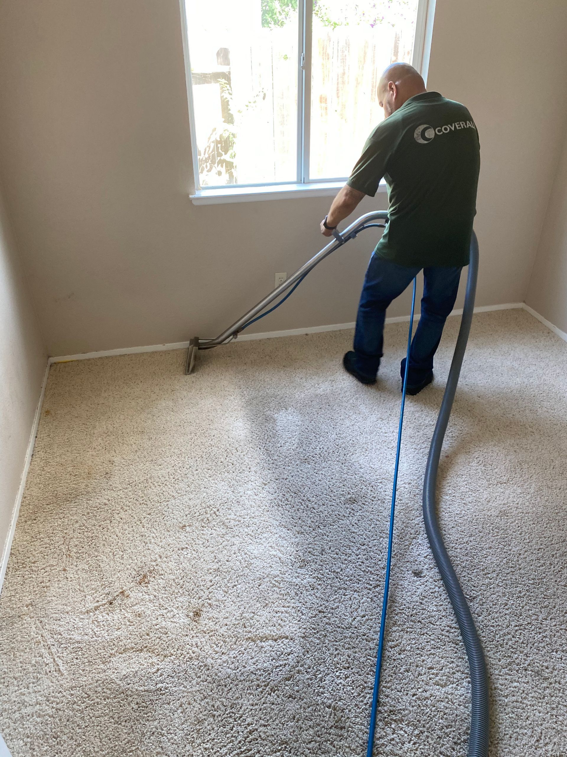 A person in a green uniform uses a steam cleaner wand to clean carpet in a room with light walls and a window.