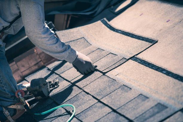 Worker repairing asphalt shingle roof with nail gun and protective gear.