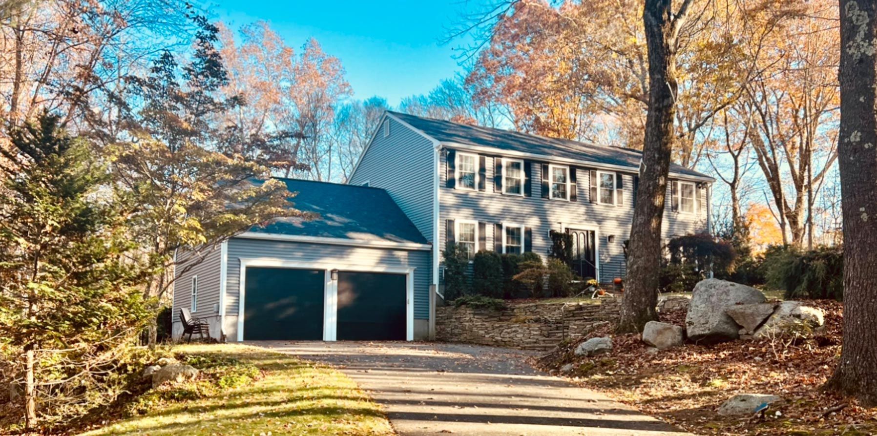 A house with two garages is surrounded by trees and leaves on a sunny day.