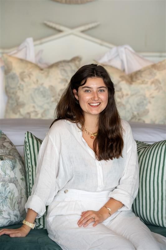Woman with blonde hair wearing a gray plaid blazer, smiling indoors near a bed and artwork.