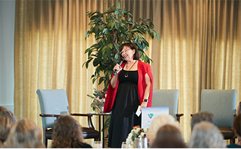 A woman in a red jacket is speaking into a microphone in front of a group of people.