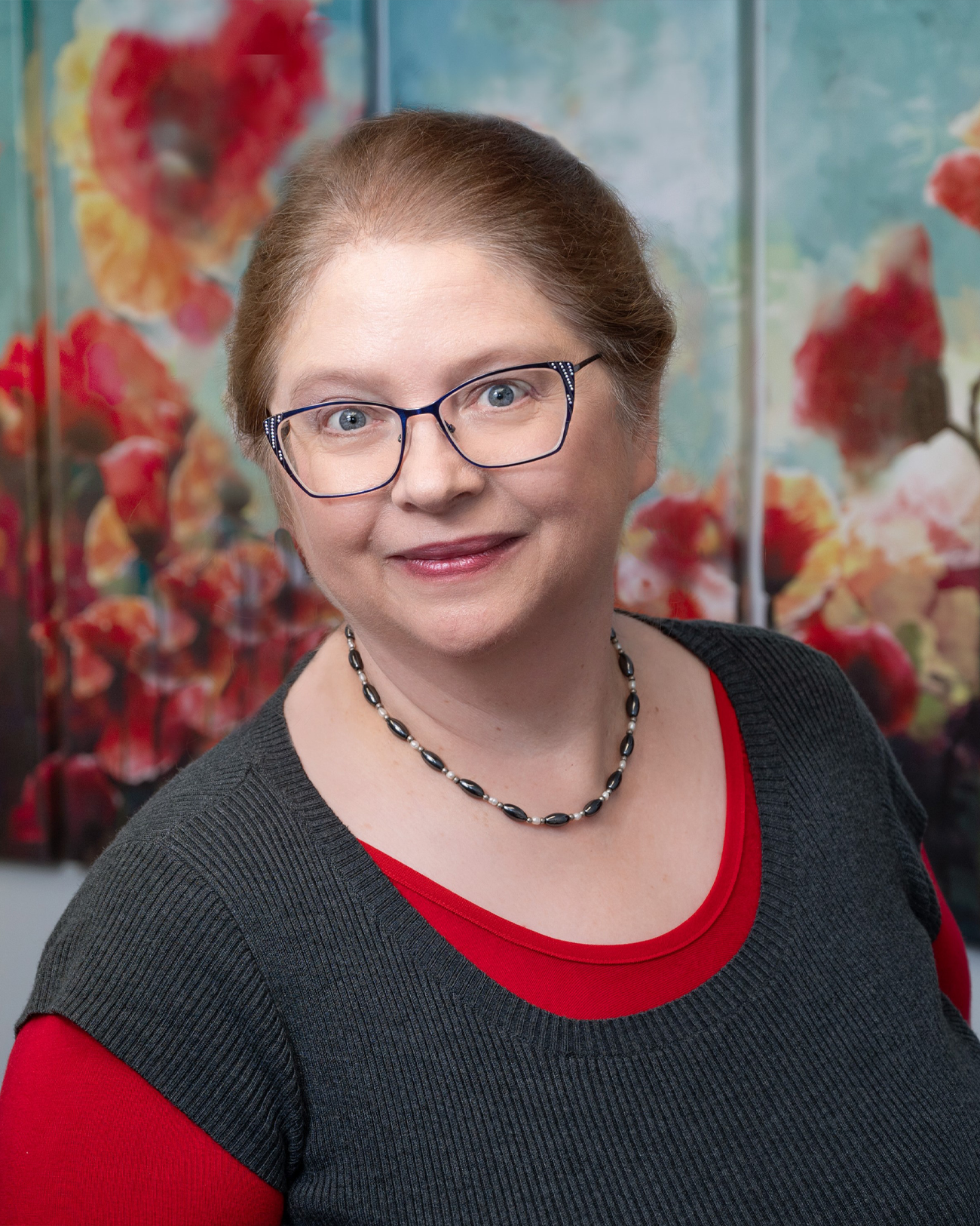 A woman wearing glasses and a necklace is standing in front of a painting of flowers.