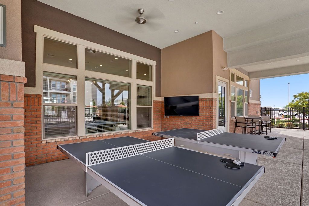 Outdoor community recreation area with two ping-pong tables, a wall-mounted TV, and seating under a covered patio.