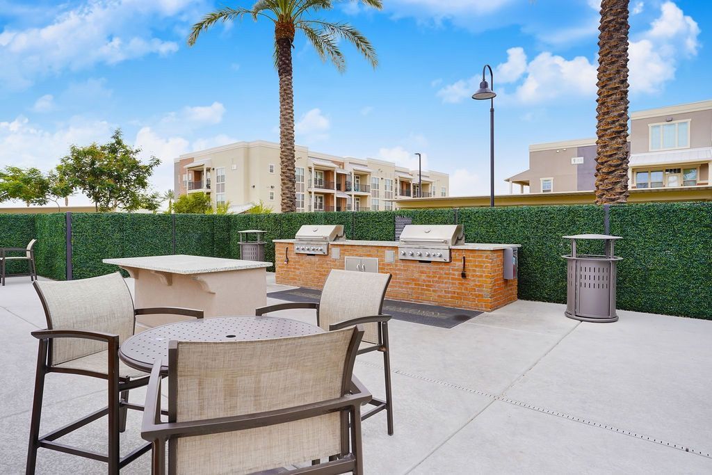 Outdoor community grilling area with tables and chairs, palm trees, and apartment buildings in the background.