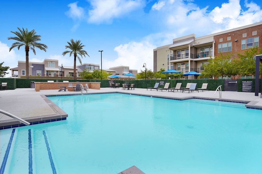 Outdoor apartment community pool with lounge chairs and blue umbrellas, surrounded by modern buildings.