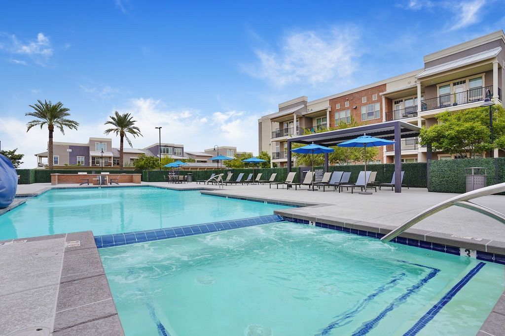 Outdoor pool with lounge chairs, blue umbrellas, and surrounding apartment buildings.