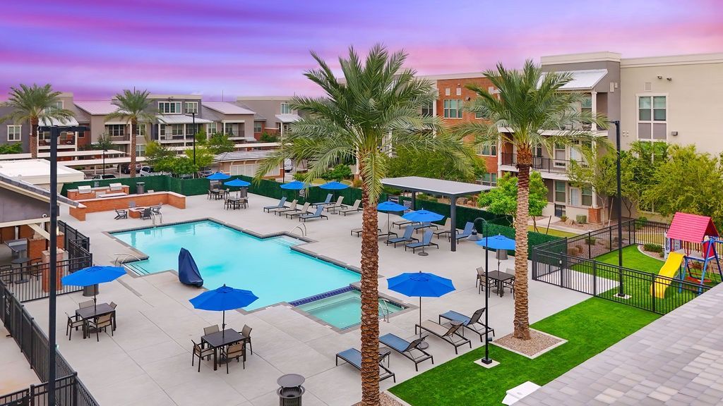 Outdoor apartment pool area with lounge chairs, blue umbrellas, and palm trees.