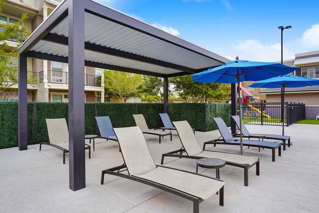Outdoor community lounge area with chaise lounges under a shaded structure and blue umbrellas.