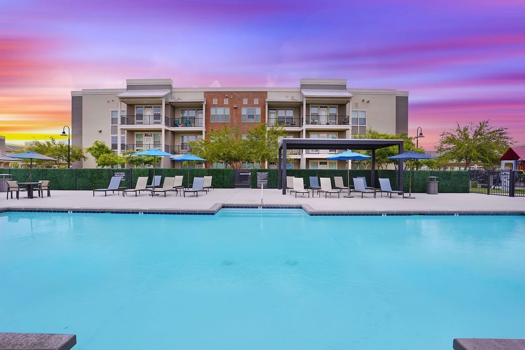 Outdoor pool area with lounge chairs and blue umbrellas beside an apartment building.