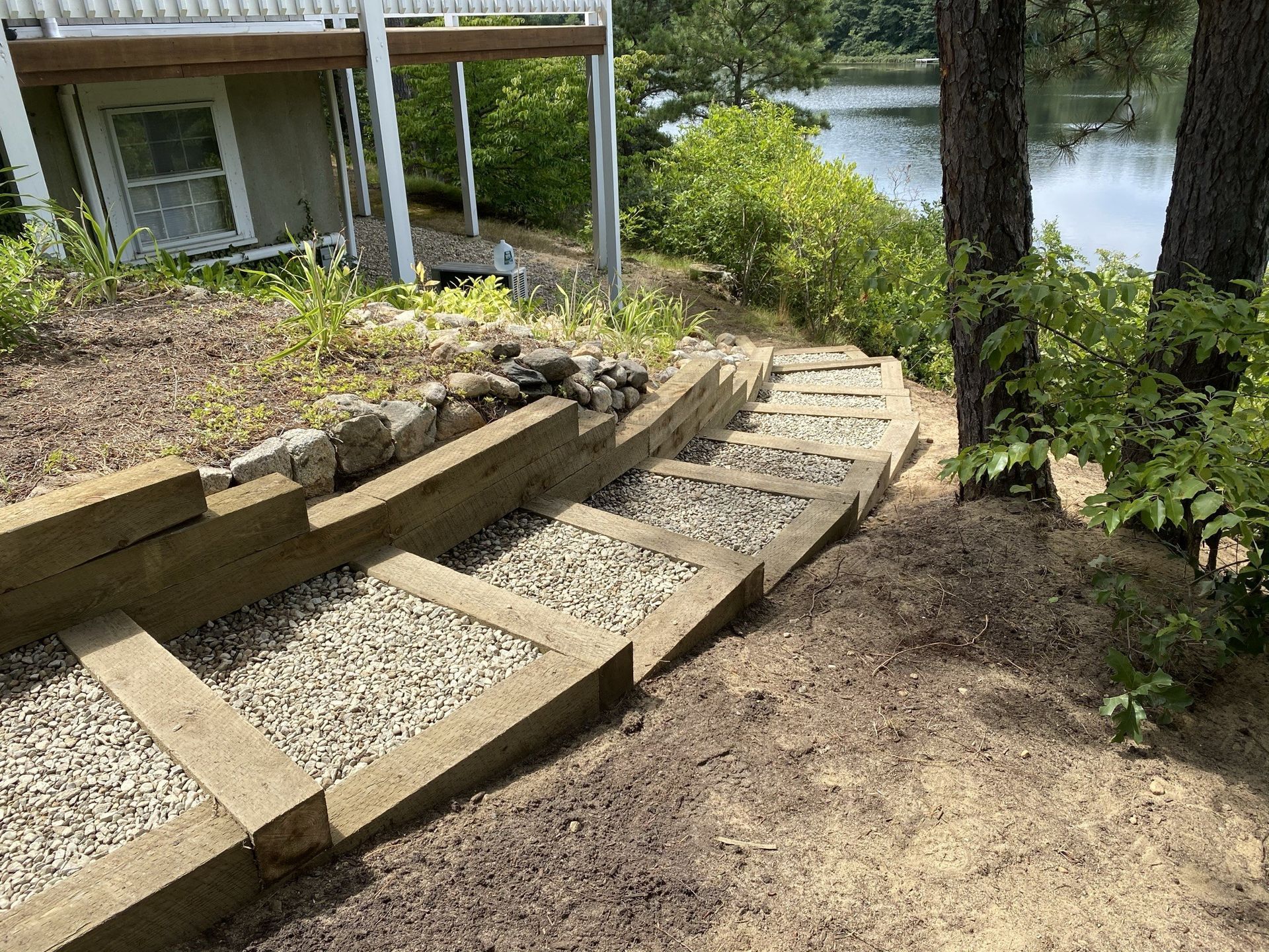 Wooden stairs filled with gravel leading down a sloped, wooded path toward a lake near a house deck.