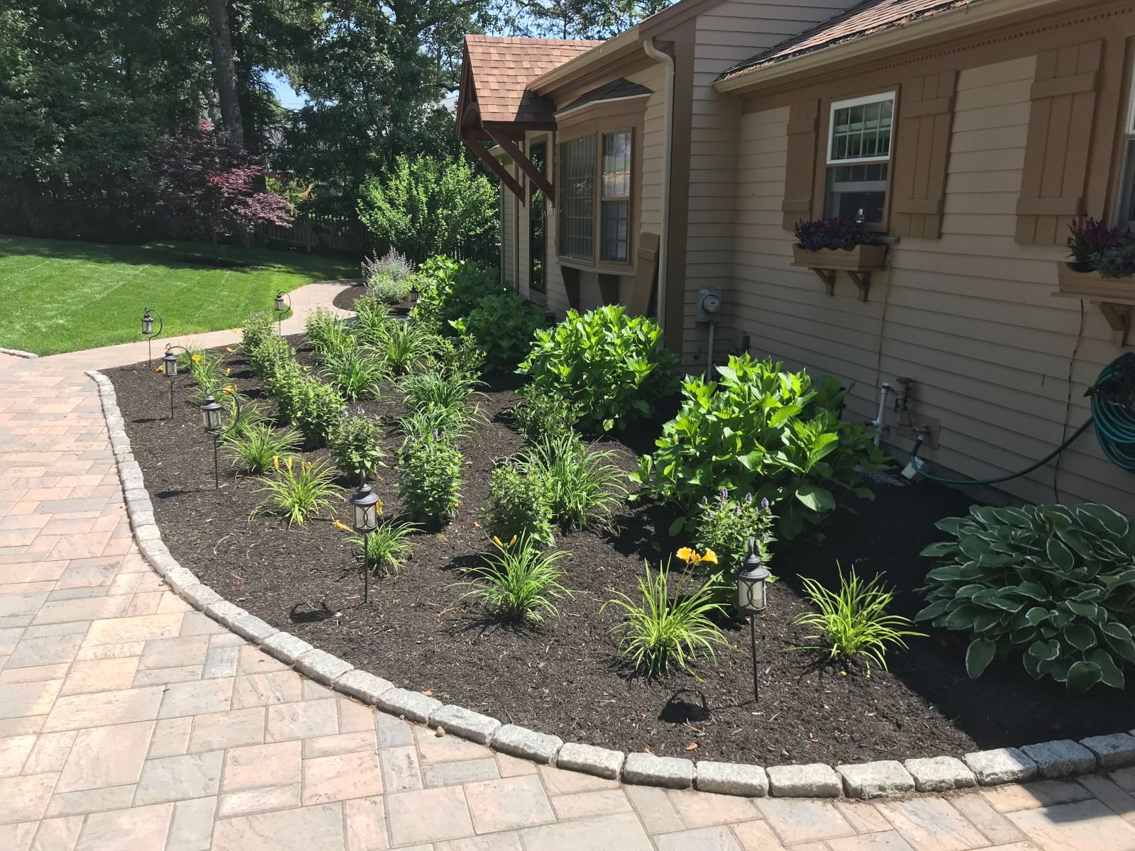 A landscaped flower bed with green shrubs and plants, edged with gray stones, next to a tan house and a brick patio.