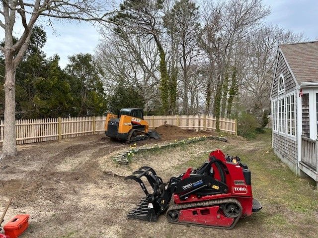 A red Toro and a yellow Case compact track loader parked in a residential yard near a wooden fence and a shingled house.