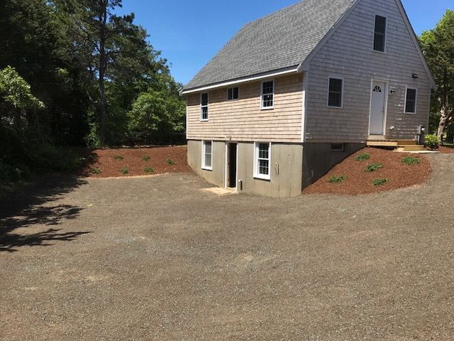 A two-story light-gray house with a daylight basement, surrounded by fresh mulch and a gravel driveway under a clear sky.