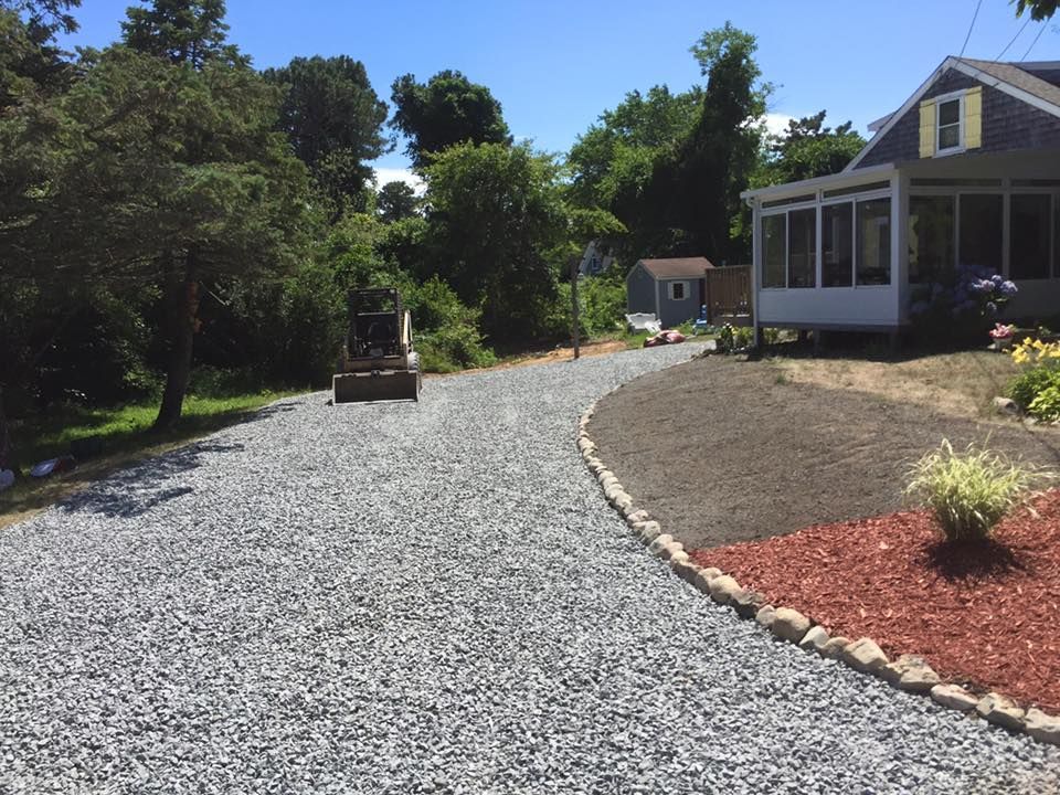 A freshly laid gravel driveway curves past a house with a screened porch, bordered by stones and a mulch garden bed.