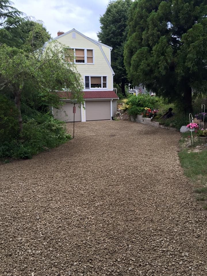 A long gravel driveway leads to a two-car garage attached to a pale yellow two-story house surrounded by trees and shrubs.
