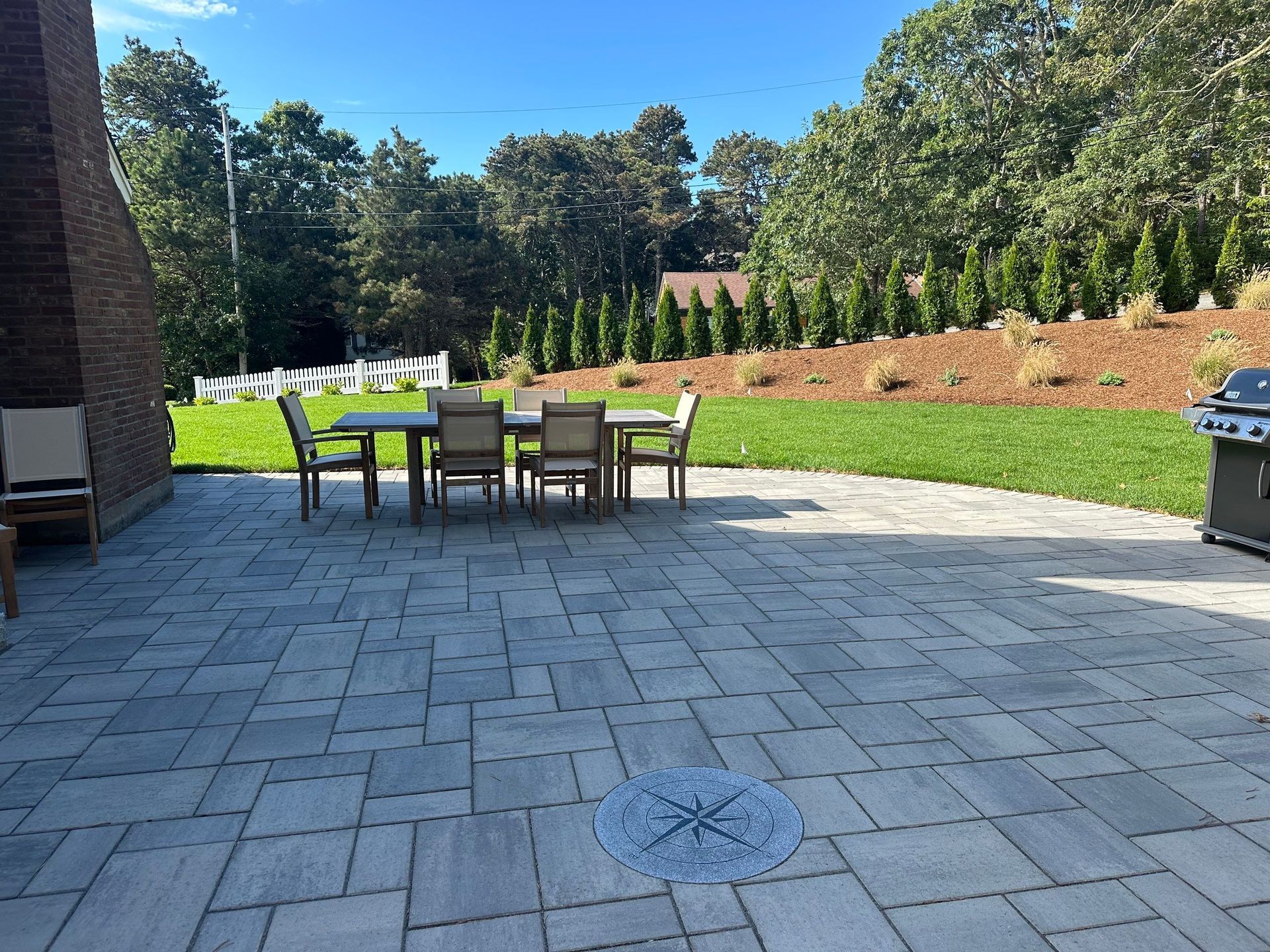 A stone patio with a dining set and a compass inlay, located in a backyard with a lawn, trees, and a brick chimney.