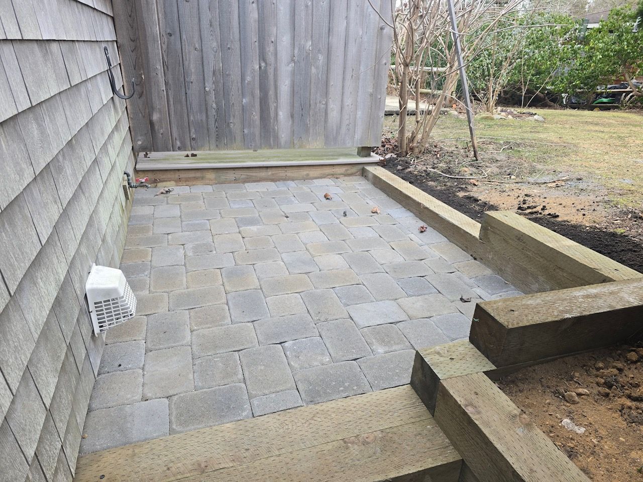 A grey stone paver patio area bordered by wooden retaining walls next to a house siding exterior.