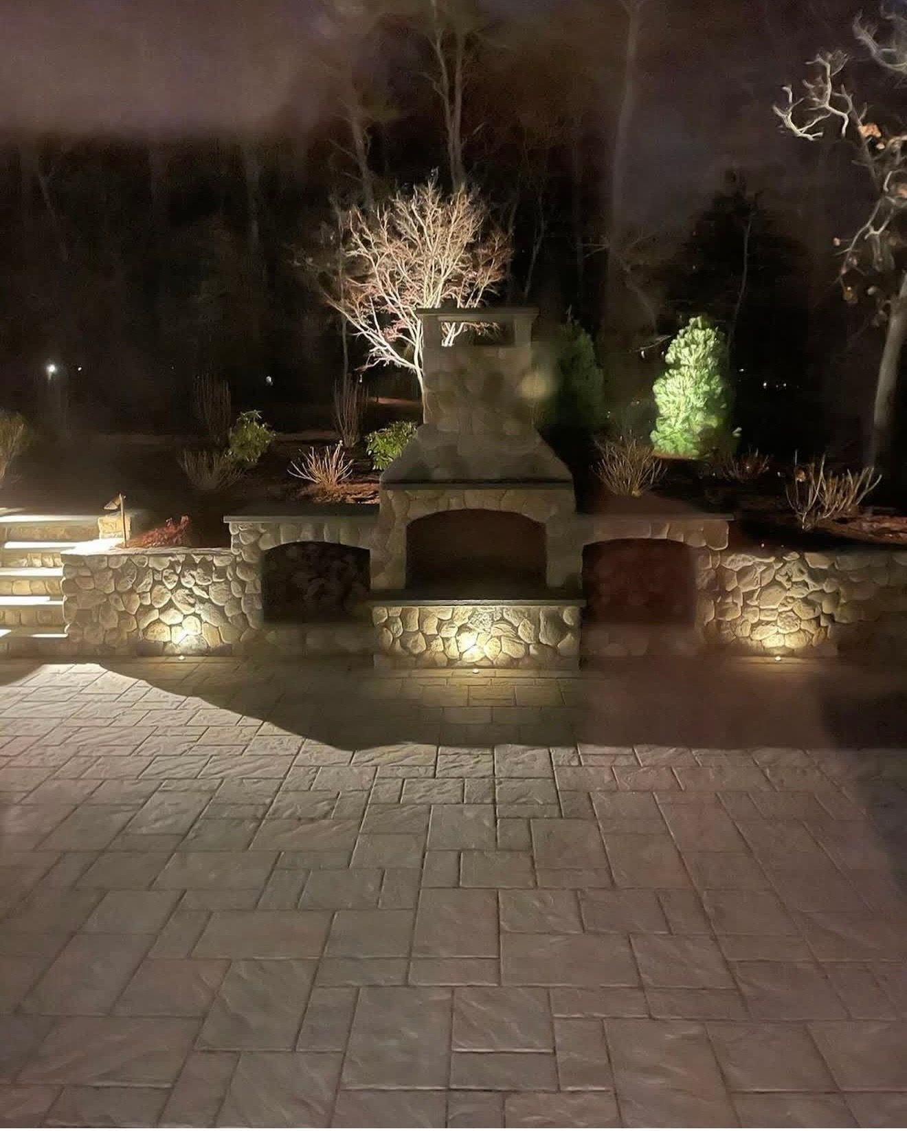 A stone outdoor fireplace and patio illuminated by landscape lighting at night, surrounded by trees and shrubbery.