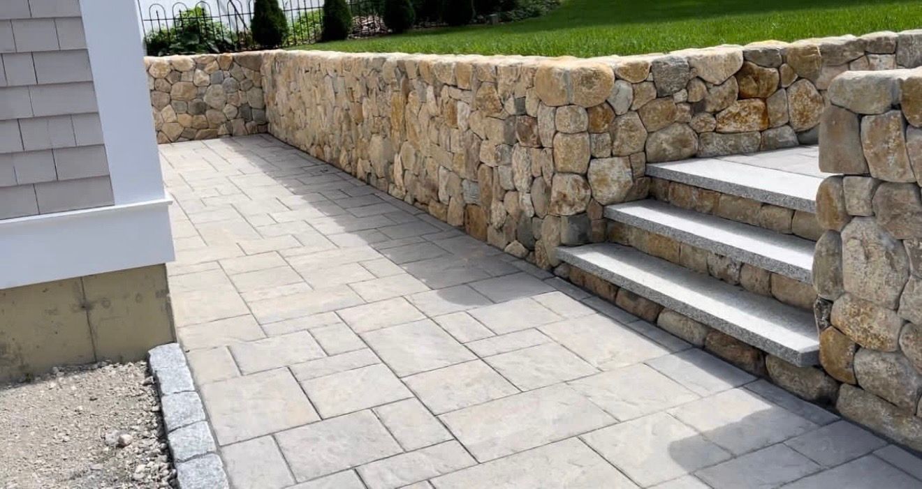 Stone retaining wall with steps leading to a green lawn, beside a paved walkway and part of a shingled building.