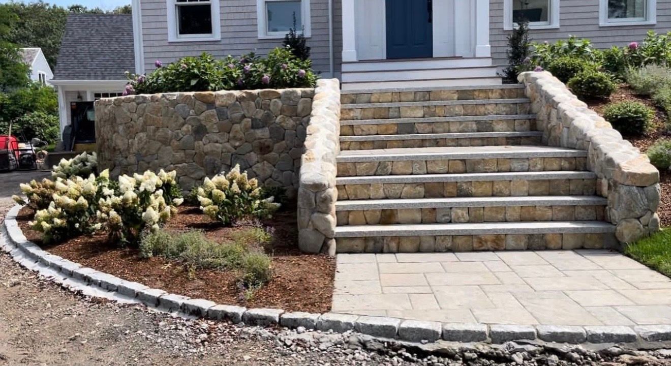 Stone steps leading to a house entrance, flanked by a curved stone retaining wall and a landscaped garden bed.