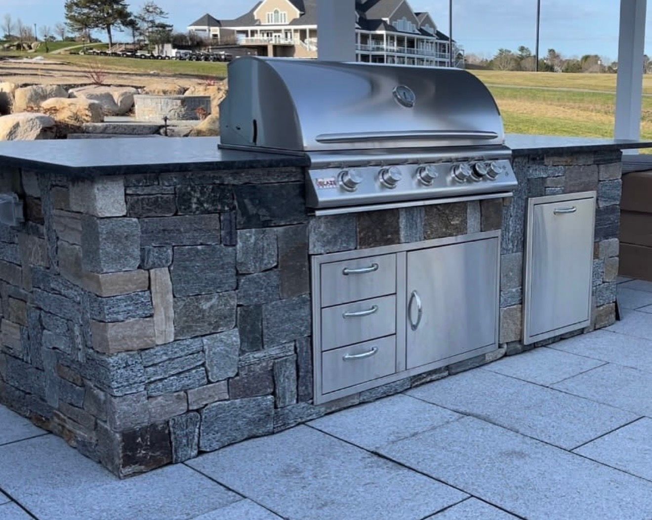 Outdoor kitchen island with stone masonry, a stainless steel gas grill, drawers, and cabinet doors on a stone patio.