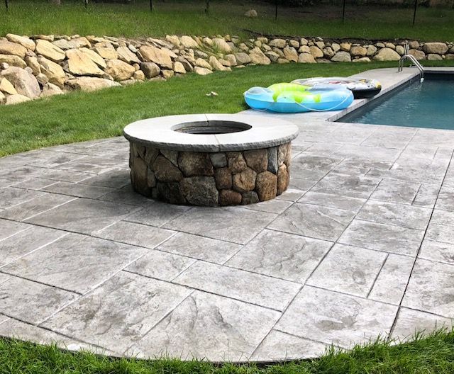 A stone fire pit on a stamped concrete patio next to a swimming pool, with a grassy area and a stone retaining wall behind.