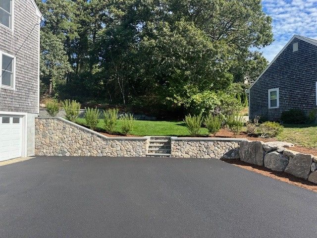 A stone retaining wall with central steps separates a paved driveway from a grassy sloped backyard between two houses.