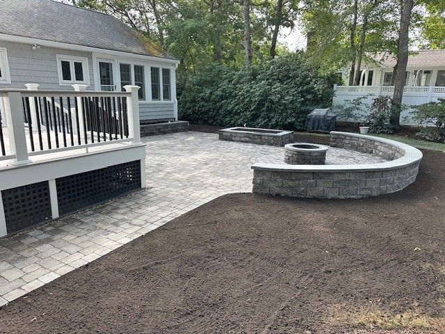 Gray patio with a curved stone fire pit wall and deck, set against a house and lush greenery.