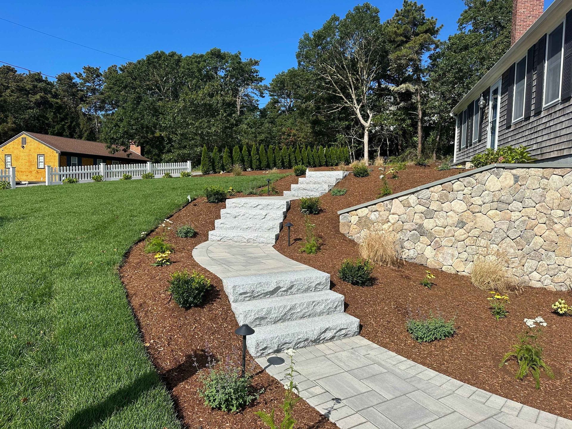 Granite stone steps and a paved walkway wind through a mulched garden beside a house and a stone retaining wall.