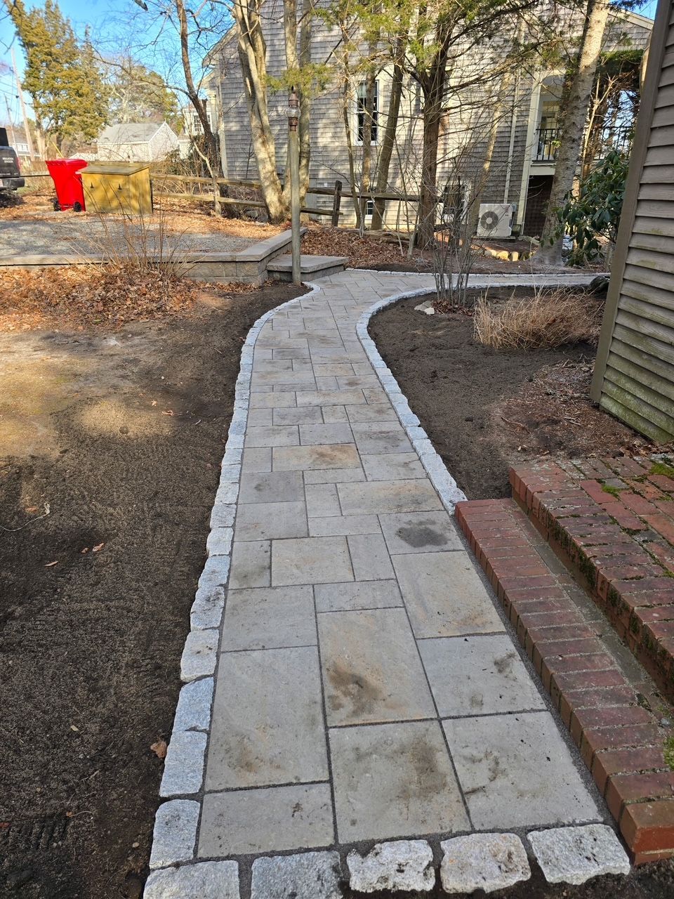 A stone paver walkway with a cobblestone border leads toward a house with brick steps, surrounded by trees and bare ground.