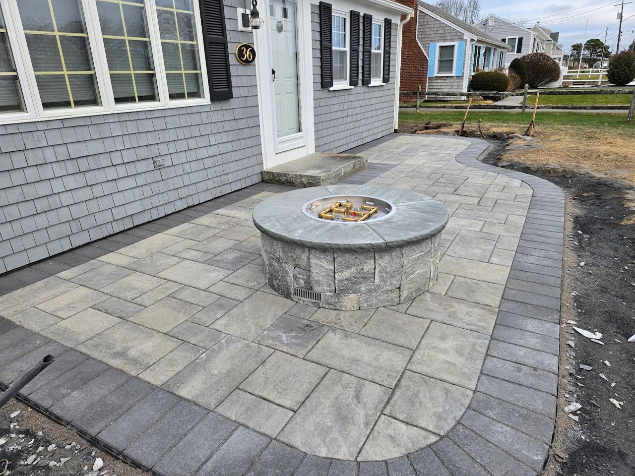 A gray house exterior with a new stone paver patio featuring a central circular fire pit and a dark border.
