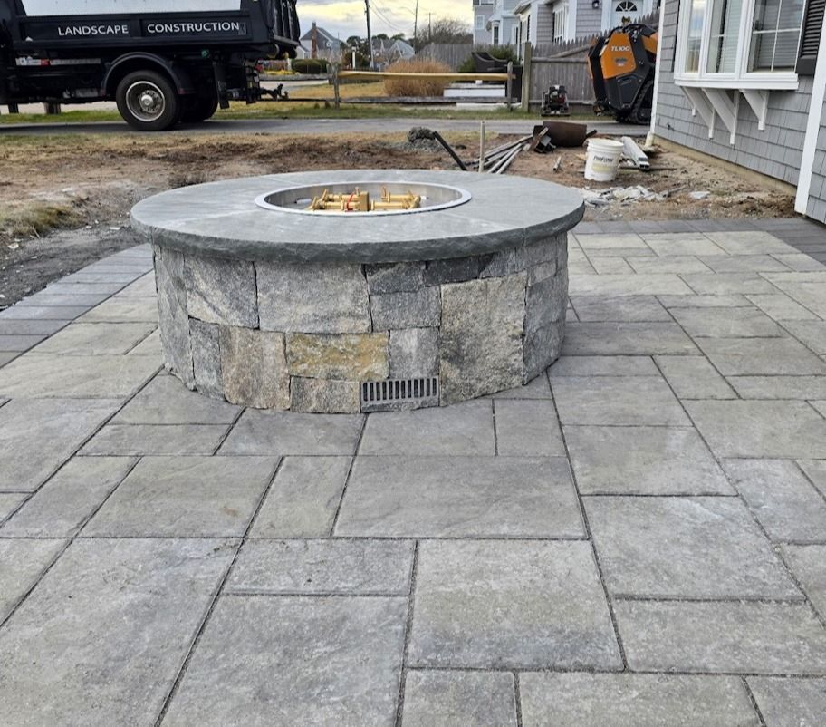 A circular stone fire pit sits on a gray patio paver surface in a backyard, with a construction truck visible in the rear.