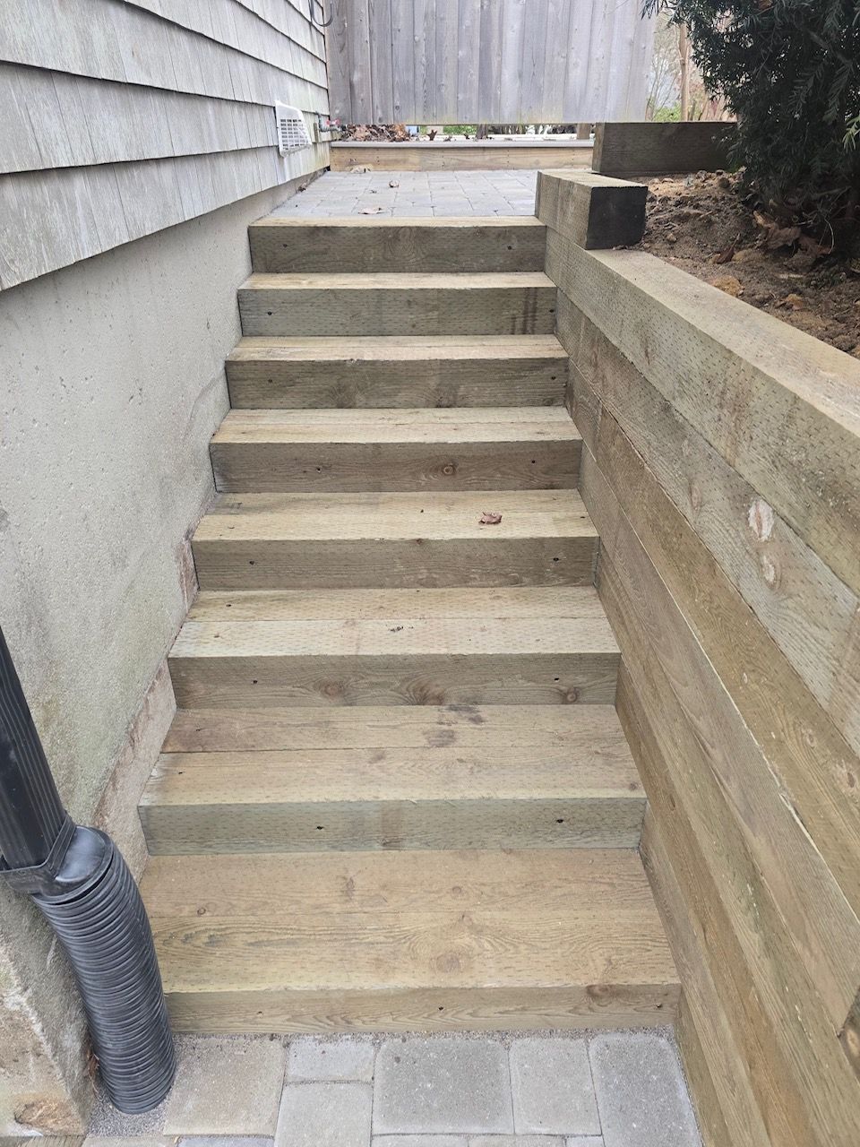 A straight flight of seven wooden steps leads up between a house wall and a low retaining wall to a paved patio area.