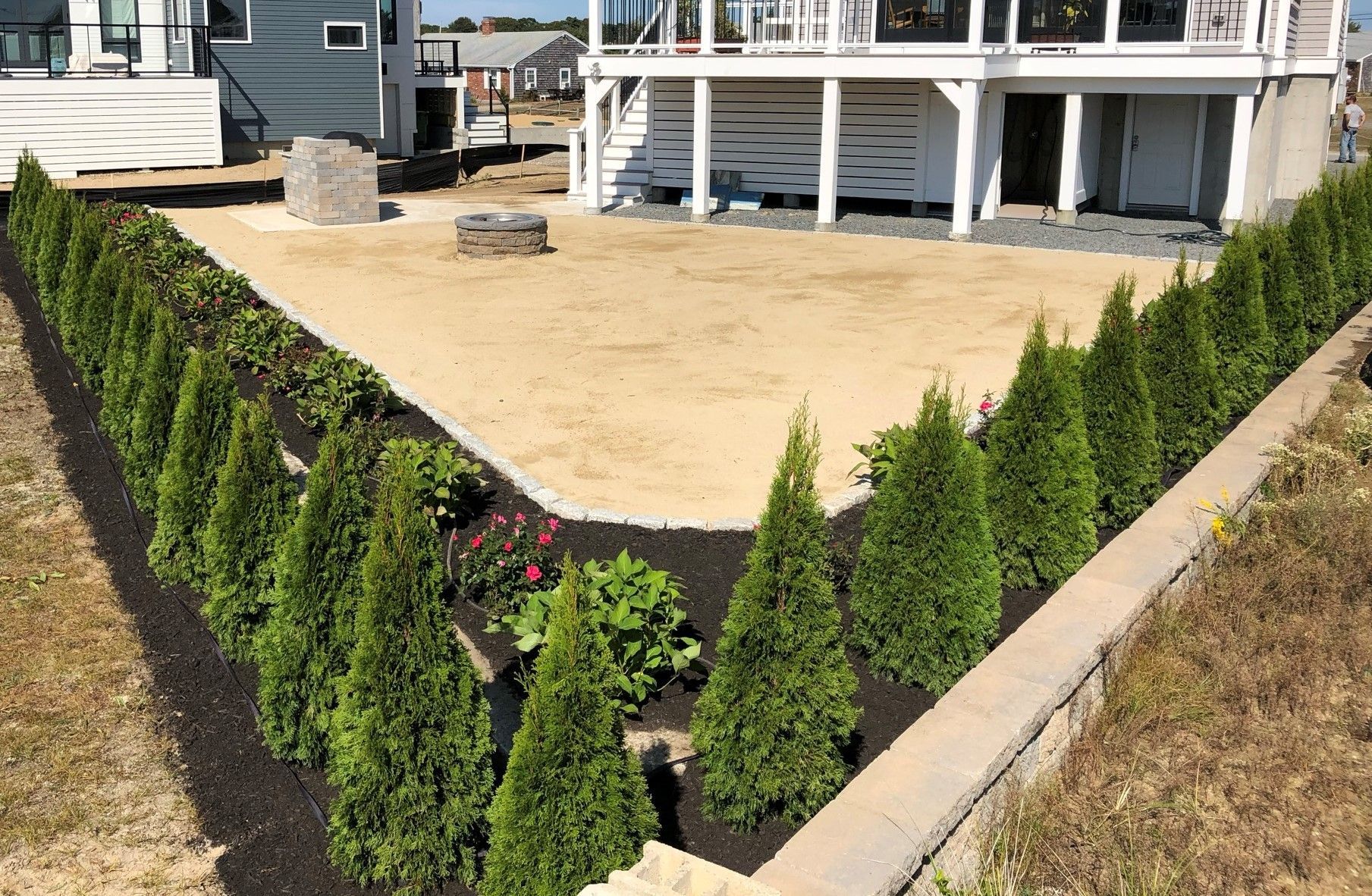 A backyard features a central sand area enclosed by a stone border, surrounded by rows of small evergreen trees and mulch.