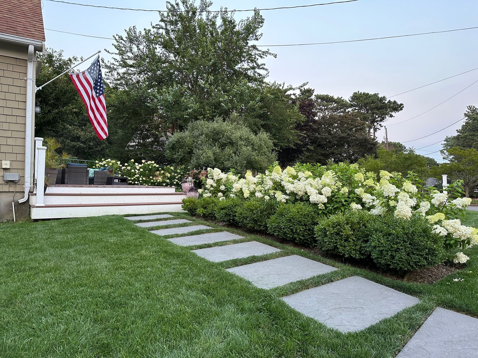 A stone path leads to a wooden deck beside a house with an American flag, bordered by white hydrangeas and green bushes.