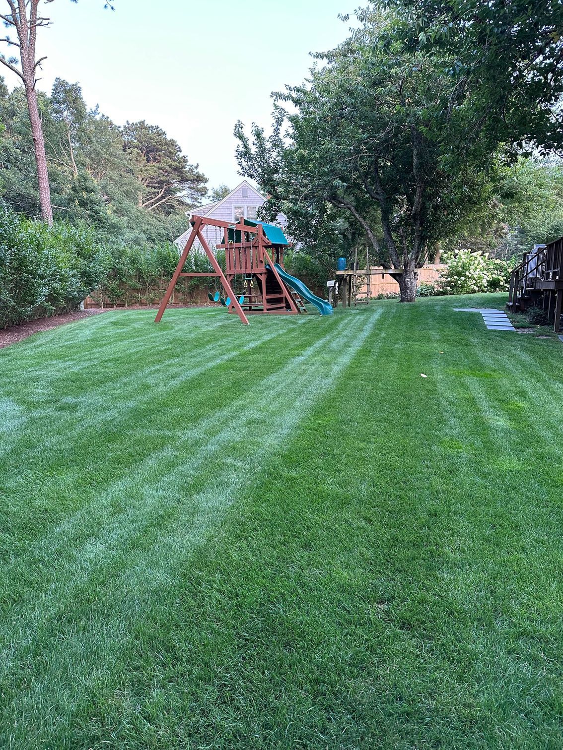 A large, well-manicured green lawn with a wooden backyard playset and slide in the distance, surrounded by trees.