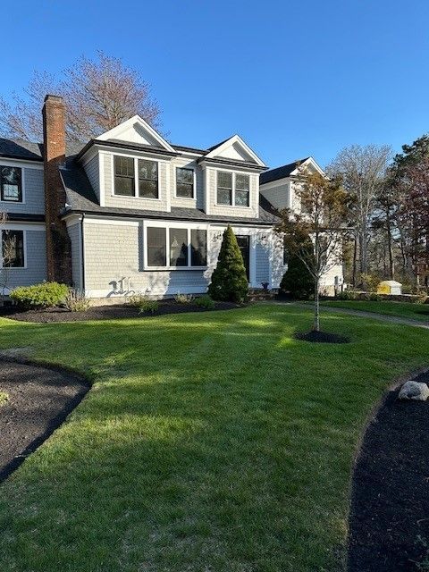 Two-story house with gray shingle siding, white trim, and a brick chimney under a clear blue sky.