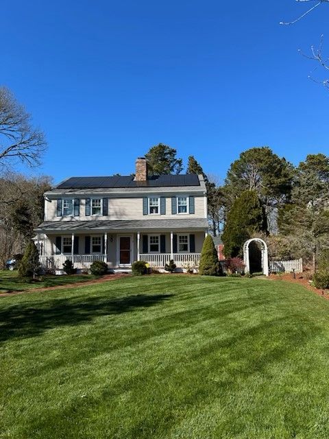 A two-story colonial house with a porch, chimney, and dark shutters, set against a lawn and blue sky.