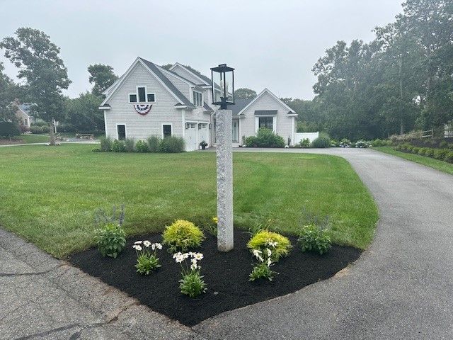 A modern suburban home with a paved driveway and a manicured lawn features a lamp post surrounded by a landscaped garden.