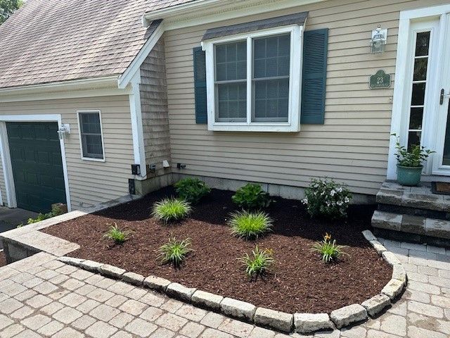 A beige house exterior with a green garage door, a mulched garden bed, and a stone paver walkway in front.