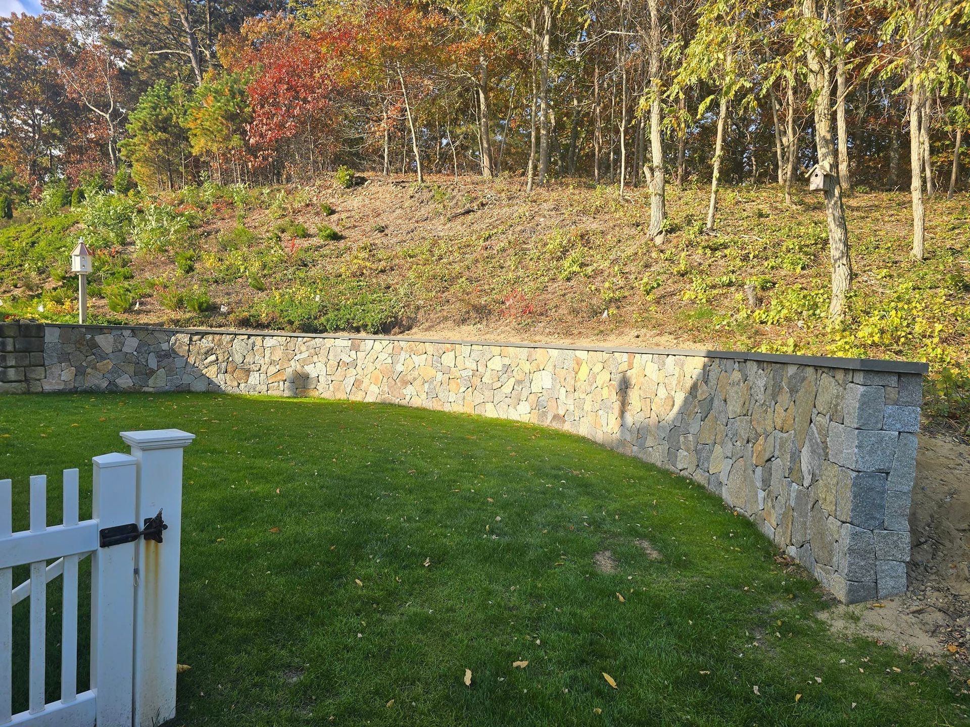 A white picket fence surrounds a lush green lawn with a stone wall in the background.