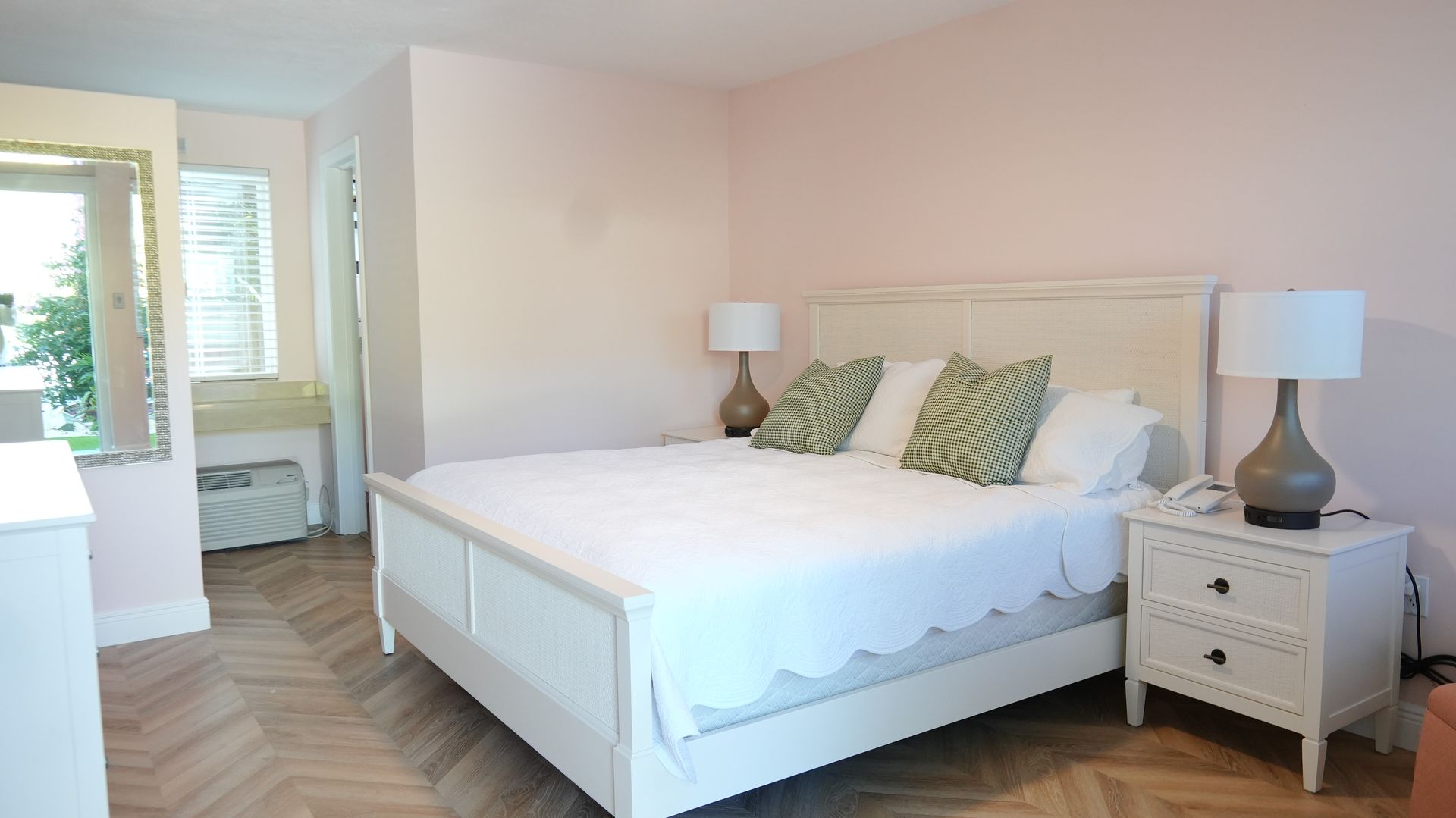 Bedroom with white bed, nightstand, lamps, and pink walls. Herringbone wood floor.